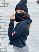 Load image into Gallery viewer, Woman wearing black crop women&#39;s hoodie with embroidered symbol on the left arm, and small red heart embroidered on the hood. Black wool beanie with Heart-OMS logo embroidered on the front.
