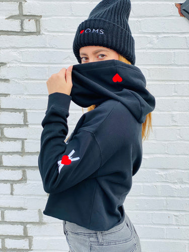 Woman wearing black crop women's hoodie with embroidered symbol on the left arm, and small red heart embroidered on the hood. Black wool beanie with Heart-OMS logo embroidered on the front.