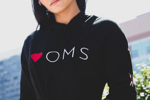 Woman wearing black crop hoodie with embroidered heart-oms symbol on the left arm and logo embroidered on the front.