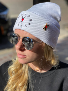 Woman wearing a white knit beanie with the circle of inspiration symbol embroidered on the fron and a 1" enamel pin of the Heart-OMS symbol pinned to the side.