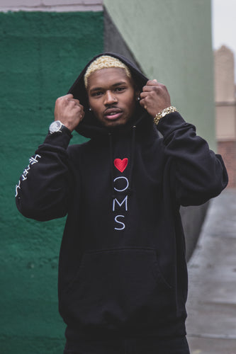 Man wearing black hoodie with heart-oms logo vertically embroidered down the front, and 