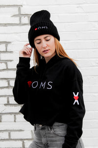 Woman wearing black crop women's hoodie with embroidered symbol on the left arm, and small red heart embroidered on the hood. Black wool beanie with Heart-OMS logo embroidered on the front.